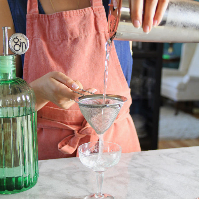 a woman pouring a martini from a cocktail tin into a stemmed glass